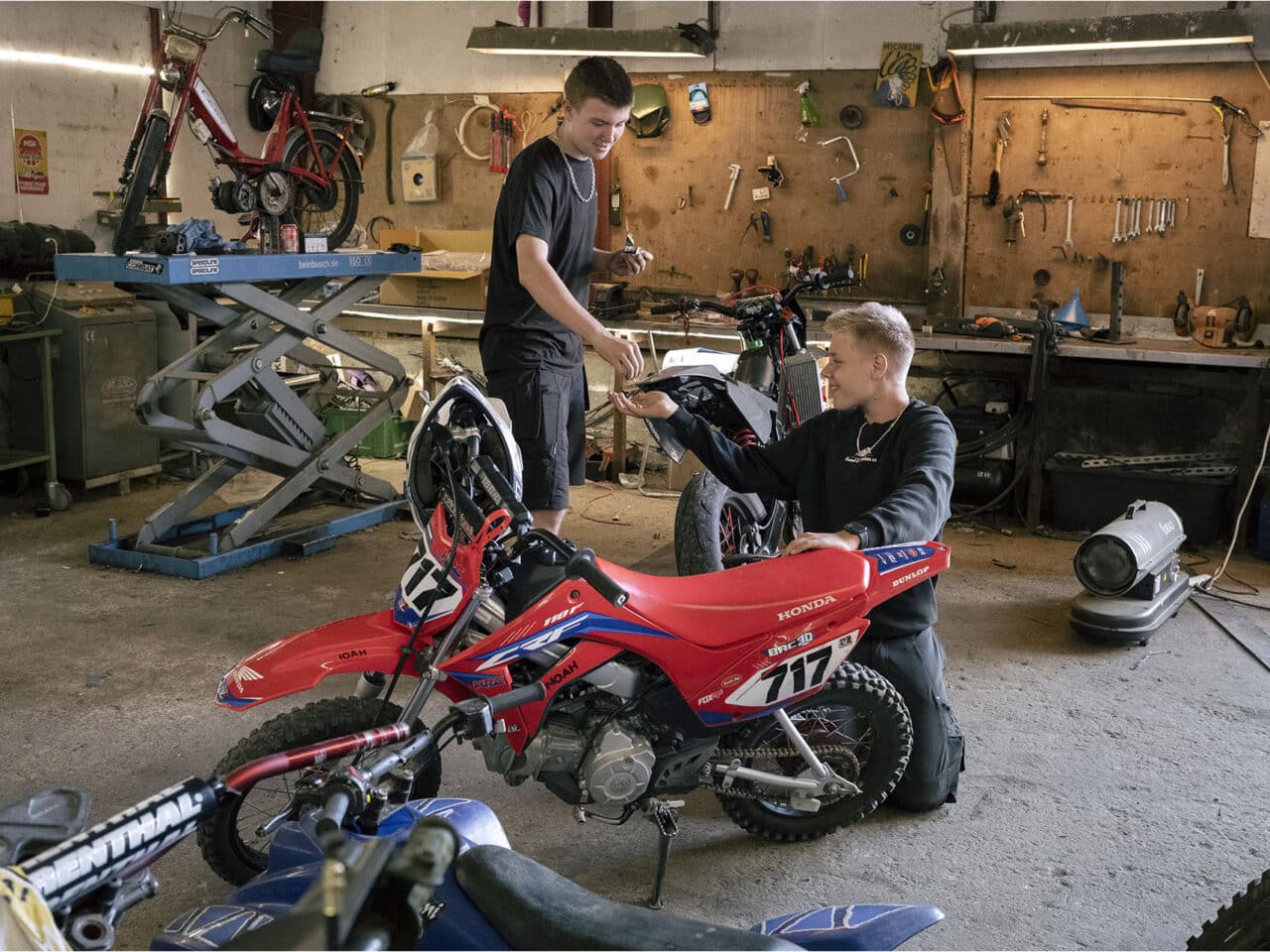 To unge står og arbejder på crossmotorcykler i et værksted med værktøj og flere motorcykler omkring sig