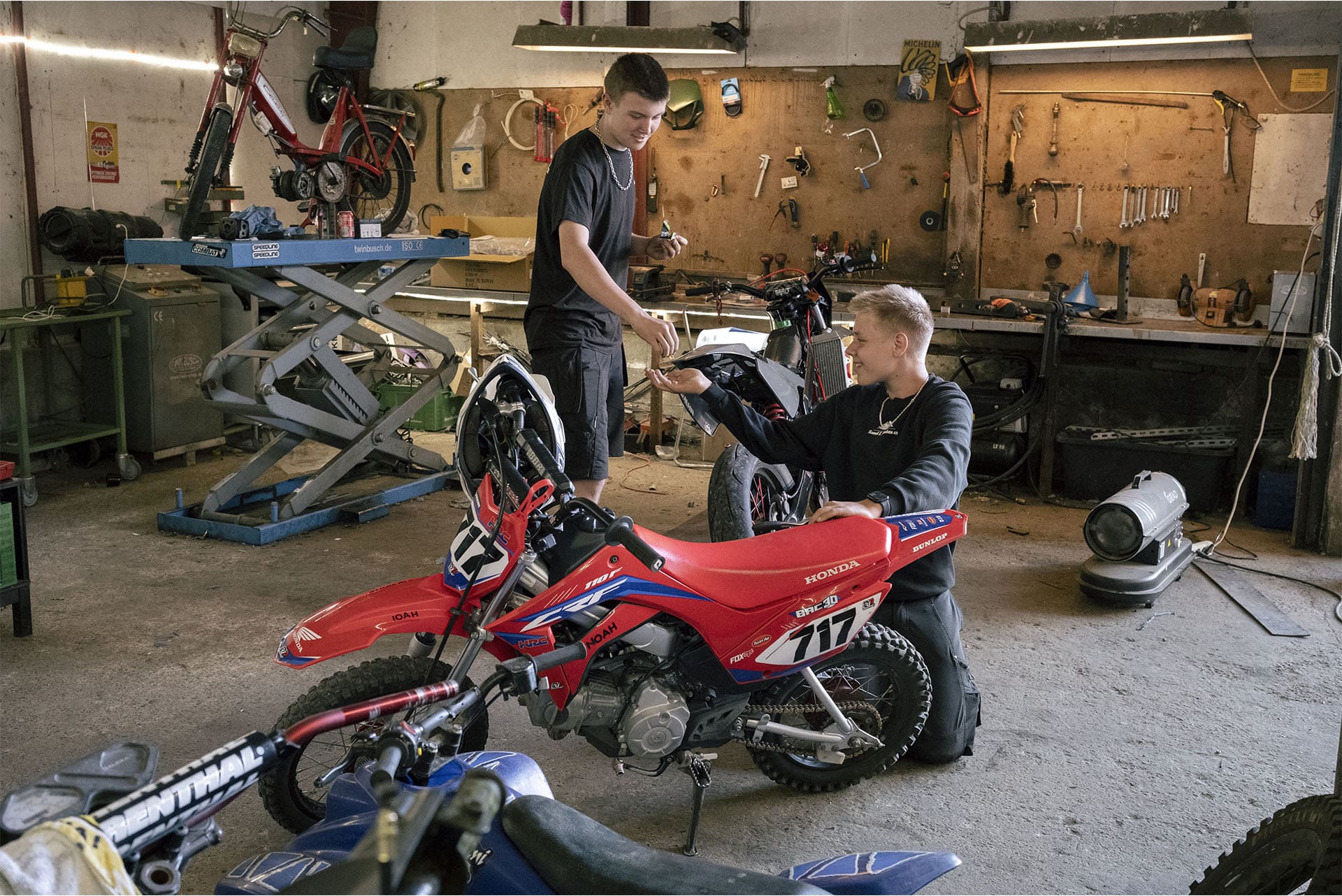 To unge står og arbejder på crossmotorcykler i et værksted med værktøj og flere motorcykler omkring sig